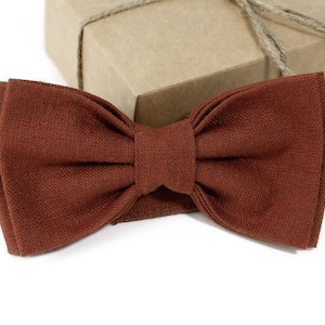 Puede incluir: Una pajarita de color óxido hecha de tela texturizada, sobre una superficie blanca. Una caja de regalo marrón con hilo de bramante está en el fondo. La pajarita es un accesorio clásico, adecuado para ocasiones formales o semiformales.