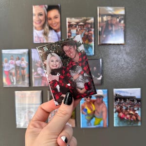 May include: A close-up of a photo magnet featuring a couple and a cat. The couple is wearing red and black plaid shirts and the cat is white and gray. The photo is held in a hand with black and white nail polish.