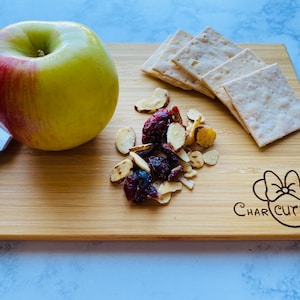 Puede incluir: Una tabla de cortar de madera con un diseño de Mickey Mouse grabado en ella. La tabla tiene una manzana roja, galletas, almendras y arándanos.