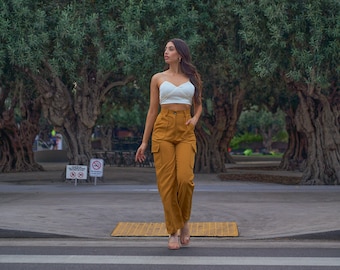 May include: A woman wearing a white crop top and mustard yellow cargo pants walks across a crosswalk in front of a row of olive trees.