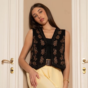 Puede incluir: Chaleco negro con bordados florales marrones y detalles de tachuelas. Se lleva sobre una camiseta negra y combinado con pantalones amarillos. Un cinturón marrón y una pulsera dorada completan el conjunto.