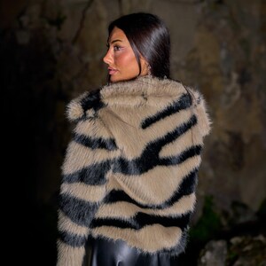 May include: A faux fur jacket with a zebra stripe pattern in black and tan. The jacket has a collar and is worn over a black garment. The image is taken in front of a stone wall.