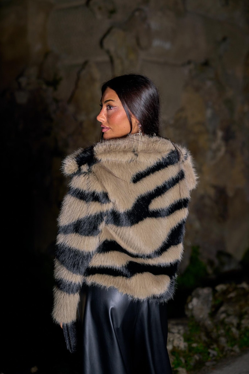 May include: A faux fur jacket with a zebra stripe pattern in black and tan. The jacket has a collar and is worn over a black garment. The image is taken in front of a stone wall.