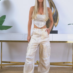 May include: A matching two-piece set featuring a metallic gold corset top and cargo pants. The pants have multiple pockets and a wide-leg design. The outfit is paired with white sneakers. The background includes a white wall and a gold-accented table.