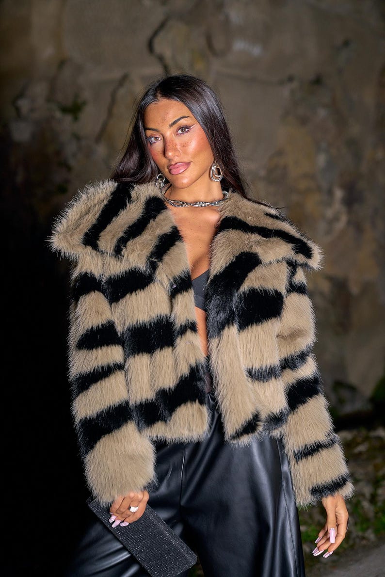 May include: A faux fur coat with a bold pattern of black stripes on a tan background. The coat has a wide collar and long sleeves. The model is wearing black leather pants and holding a black clutch.