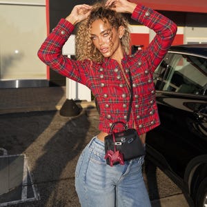 May include: A red plaid military-style jacket featuring long sleeves, a high collar, and two vertical rows of silver-toned metallic buttons. The jacket is paired with light blue denim jeans and a small black structured handbag with a red charm.
