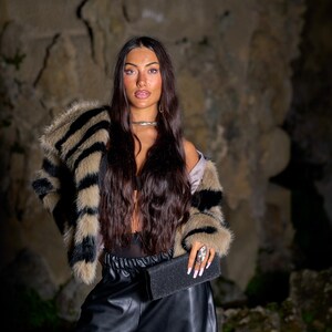 May include: A woman wearing a faux fur jacket with black and tan stripes, black leather pants, and a black clutch. The jacket has a wide collar and cuffs. The clutch is rectangular and textured. The background is a stone wall.