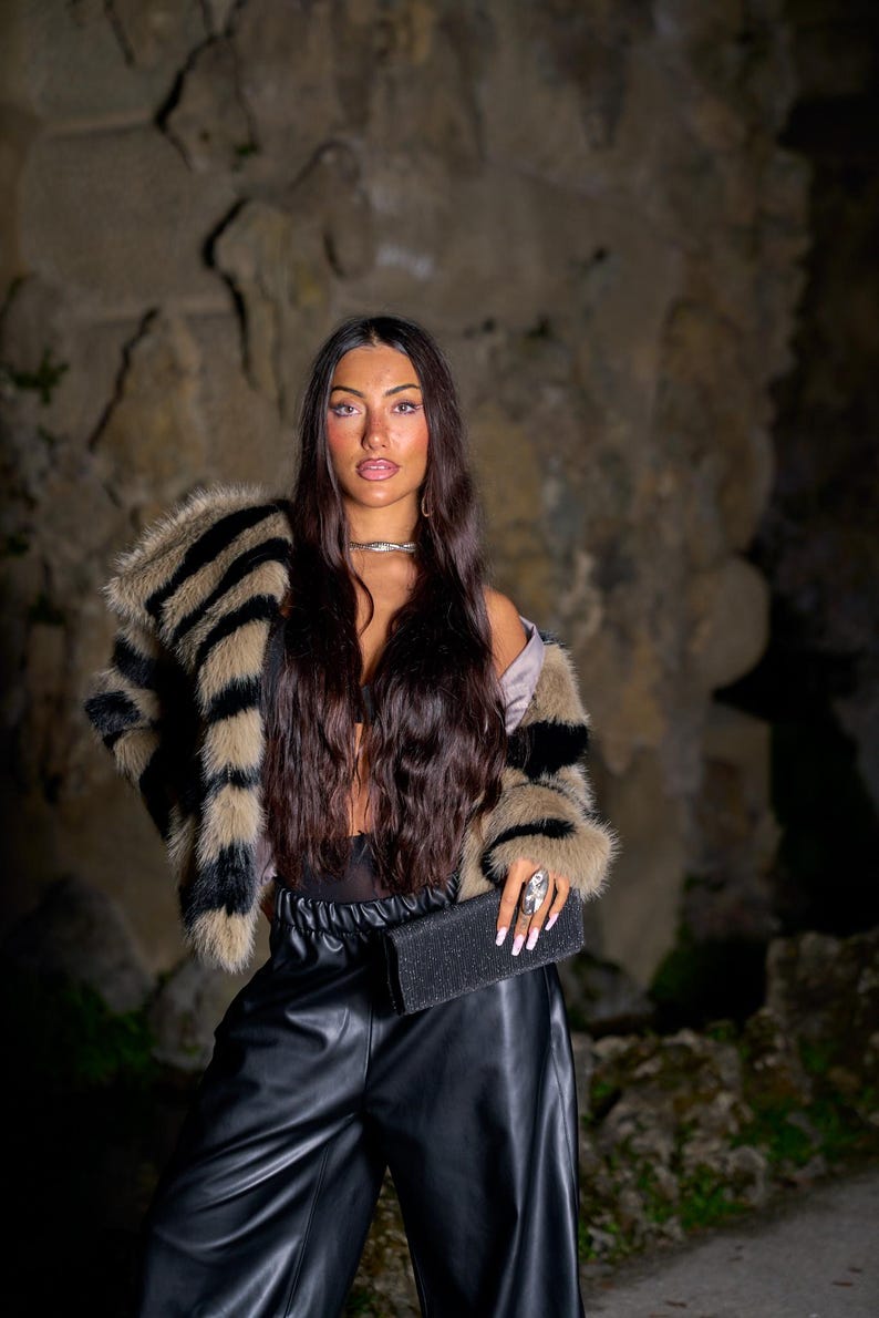 May include: A woman wearing a faux fur jacket with black and tan stripes, black leather pants, and a black clutch. The jacket has a wide collar and cuffs. The clutch is rectangular and textured. The background is a stone wall.
