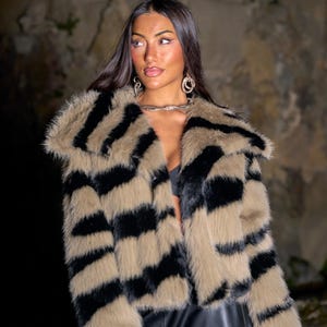 May include: A faux fur jacket with a striking zebra stripe pattern in black and tan. The jacket has a wide collar and long sleeves. The model is wearing a black top and black leather pants. The background is a textured, neutral color.