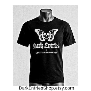 Può includere: T-shirt nera con grafica bianca raffigurante una farfalla con un teschio al centro, le parole "Dark Entries" e "USCITE DI SICUREZZA" in bianco. La maglietta è esposta su un manichino.