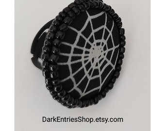May include: A circular ring featuring a black fabric center with a grey spiderweb pattern. The edge of the circular face is decorated with a border of small, shiny black beads. The text DarkEntriesShop.etsy.com is printed below the ring.