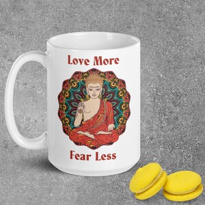 love more fear less coffee mug with handle by goddess swag. colorful buddha image with mandala behind it. Love More is written in a deep red above the buddha image and fear less is written in a deep red below the buddha image. Double sided.