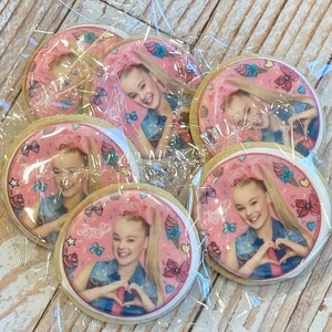 May include: A set of six round sugar cookies decorated with a pink background and an image of a young girl with blonde hair. The girl is wearing a denim jacket and has a heart-shaped hand gesture. The cookies are packaged in clear plastic bags.