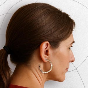 May include: A woman wearing gold-colored hoop earrings. The earrings have a textured, organic design. The woman is wearing a red shirt and has her hair pulled back in a ponytail. The background is white with gray lines.