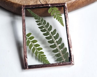 May include: A copper framed pendant with a pressed fern inside. The pendant is on a wooden surface.