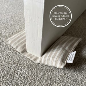 May include: A beige and white striped door wedge is positioned under a white door. The doorstop is made of fabric and has a small tag that reads "Katy Wiese Homeware". The image also includes text that says "Door Wedge Sewing Tutorial Digital PDF".