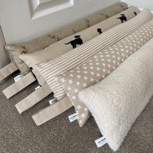 May include: Five doorstop cushions in various patterns. The cushions are made of a soft, beige fabric. The patterns include a white and beige striped pattern, a white star pattern, and a beige fabric with a brown and white animal print. The cushions have a beige fabric strap with a label that reads "Katy Walker Designs".