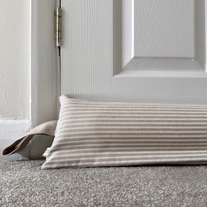 May include: A long, rectangular fabric door draught excluder featuring a pattern of thin, alternating beige and white horizontal stripes. The item is constructed with a sturdy fabric handle on the left side and is positioned horizontally along the base of a door.