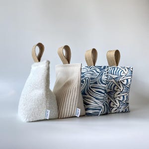 May include: Four wedge-shaped doorstops with jute fabric handles. The first is white textured fleece, the second is beige and white striped fabric, the third has a blue and white wave pattern, and the fourth features a blue and white fish print.