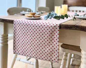 May include: A cream-colored table runner with a repeating pattern of red stars. The runner is placed on a wooden table, partially covering it. The table is set with plates, candles, and greenery, suggesting a festive or holiday theme.