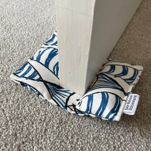 May include: A fabric doorstop featuring a white base with a blue and black abstract leaf pattern. The rectangular, stuffed object is positioned under a white door. A small white fabric tag on the side displays the text Katy Webster Homeware.