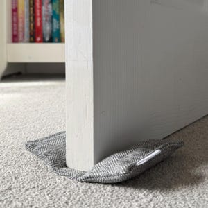 May include: A grey and white herringbone patterned fabric doorstop wedge positioned at the base of a white wooden door. The rectangular, soft-filled textile object is designed to hold the door in place, featuring a small white tag on the side.