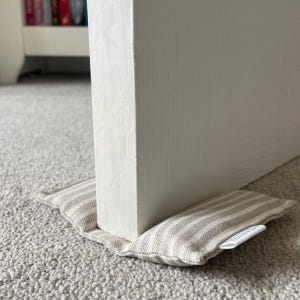 May include: A rectangular fabric doorstop featuring a beige and white striped pattern. The doorstop is positioned at the base of a white wooden door, resting on a grey carpeted floor. A small white fabric tag is visible on the side of the item.