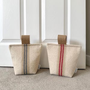 May include: Two wedge-shaped doorstops made of beige fabric with a textured weave. Each doorstop features a vertical stripe pattern and a brown fabric handle at the top. One doorstop has a blue stripe, and the other has a red stripe.