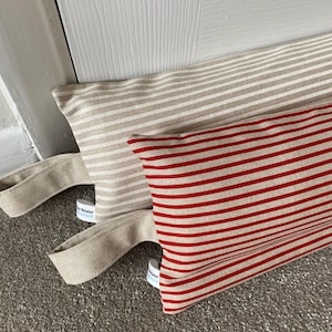 May include: Two striped doorstops, one beige and white, the other red and white. Each has a fabric handle. They are positioned on a grey carpet near a white door frame.