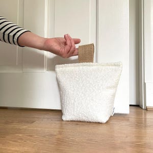 May include: A white, textured, sherpa-style fabric doorstop with a rectangular shape and a tan, woven burlap handle attached to the top. The doorstop features a small white fabric tag on the side with black text printed on it.