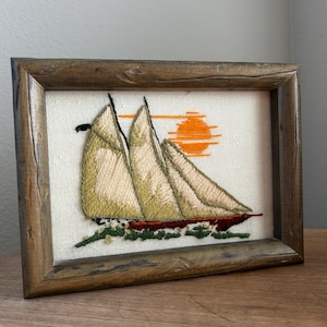 Puede incluir: Obra de arte bordada de un velero con velas beige y casco rojo, sobre un fondo crema con un sol naranja. La obra está enmarcada en un marco de madera marrón rústico.