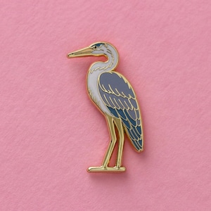 May include: A gold-colored enamel pin featuring a blue and gray heron with a long, thin beak. The heron is standing on one leg with its wings folded.