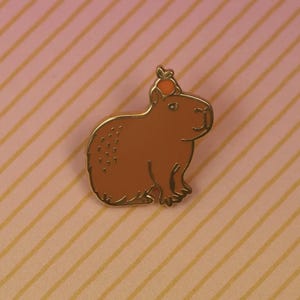 May include: A brown capybara enamel pin with gold trim, wearing a small orange fruit on its head. The pin is set against a pink background with yellow diagonal stripes. A cute animal-themed accessory.