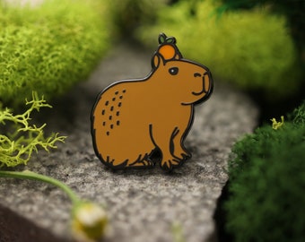 May include: A brown capybara enamel pin with a small orange fruit on its head. The pin has a black outline and is set against a green mossy background.