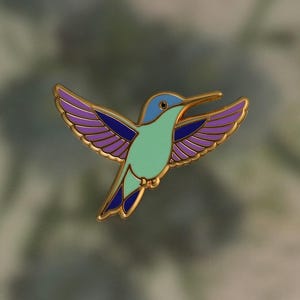 May include: A gold-toned hummingbird pin with a vibrant design. The bird features a light blue head, a mint green body, and purple wings with gold accents. The wings are spread in mid-flight.
