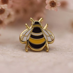 Op de afbeelding: Een gouden emaille pin met een cartoon-hommel met zwarte en gele strepen en witte vleugels.
