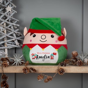 May include: A plush elf toy with a green hat and a red and white striped shirt. The elf has a friendly face and is holding a sign that says "Merry Christmas Amelia".