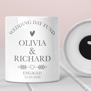 May include: White ceramic piggy bank with a slot on top. The front of the bank has the text "Wedding Day Fund" with a heart, the names "Olivia & Richard", the text "Engaged" and the date "25/07/2020".