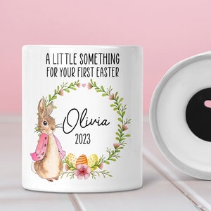 Puede incluir: Hucha de cerámica blanca con un diseño de conejo y una corona de flores. El texto dice "Un pequeño detalle para tu primera Pascua" y "Olivia 2023".