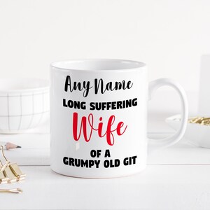 Può includere: Tazza in ceramica bianca con testo nero che recita "Any Name Long Suffering Wife of a Grumpy Old Git". La parola "Wife" è in rosso.