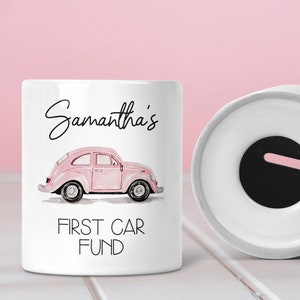 Puede incluir: Hucha de cerámica blanca con una ilustración de coche rosa y el texto "Samantha's FIRST CAR FUND". Una tapa de cerámica blanca y una pequeña ranura para monedas blanca están al lado del banco. El fondo es rosa.