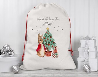 May include: A white Christmas gift bag with red drawstrings, featuring a watercolor illustration of a rabbit, a Christmas tree, and presents. The bag is personalized with the text "Special Delivery For Maisie".