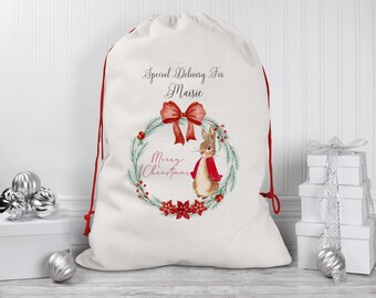 May include: A white Christmas gift bag with red drawstring, featuring a watercolor illustration of a rabbit in a red coat, a wreath with red berries, and the text "Special Delivery For Maisie Merry Christmas".