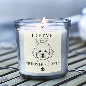 Peut inclure: Une bougie blanche dans un pot en verre transparent avec une étiquette qui dit "LIGHT ME When The BICHON FRISE FARTS" et une image de dessin animé d'un chien Bichon Frisé.