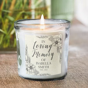 Puede incluir: Una vela blanca en un tarro de vidrio transparente con una etiqueta que dice "In Loving Memory of Isabella Smith 1941 - 2022". La etiqueta tiene un diseño floral con flores blancas y negras.