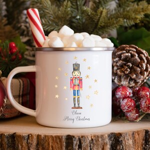 Puede incluir: Taza de esmalte blanco con una ilustración de cascanueces y estrellas doradas. La taza tiene un cascanueces rojo y azul con un sombrero negro y un borde dorado. El texto "Olivia Feliz Navidad" está impreso en la taza.
