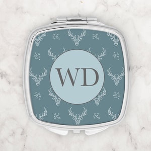 May include: A silver compact mirror with a blue background and a white geometric deer pattern. The mirror has a round, light blue center with the initials "WD" in gray.
