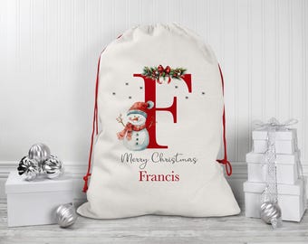 Puede incluir: Una media de Navidad blanca con un cordón de ajuste rojo y una letra roja "F" decorada con un muñeco de nieve y vegetación. La media dice "Merry Christmas Francis".