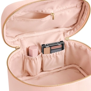 Pode incluir: Uma bolsa de maquiagem oval rosa com um z&iacute;per dourado. A bolsa est&aacute; aberta e tem v&aacute;rios compartimentos dentro. H&aacute; uma pequena paleta de sombras para os olhos e um batom rosa dentro da bolsa.
