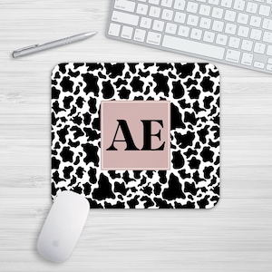 Könnte beinhalten: Ein Mauspad mit Kuhmuster in Schwarz und Weiß mit einem rosa Quadrat in der Mitte. Das Quadrat enthält die Buchstaben "AE" in Schwarz.